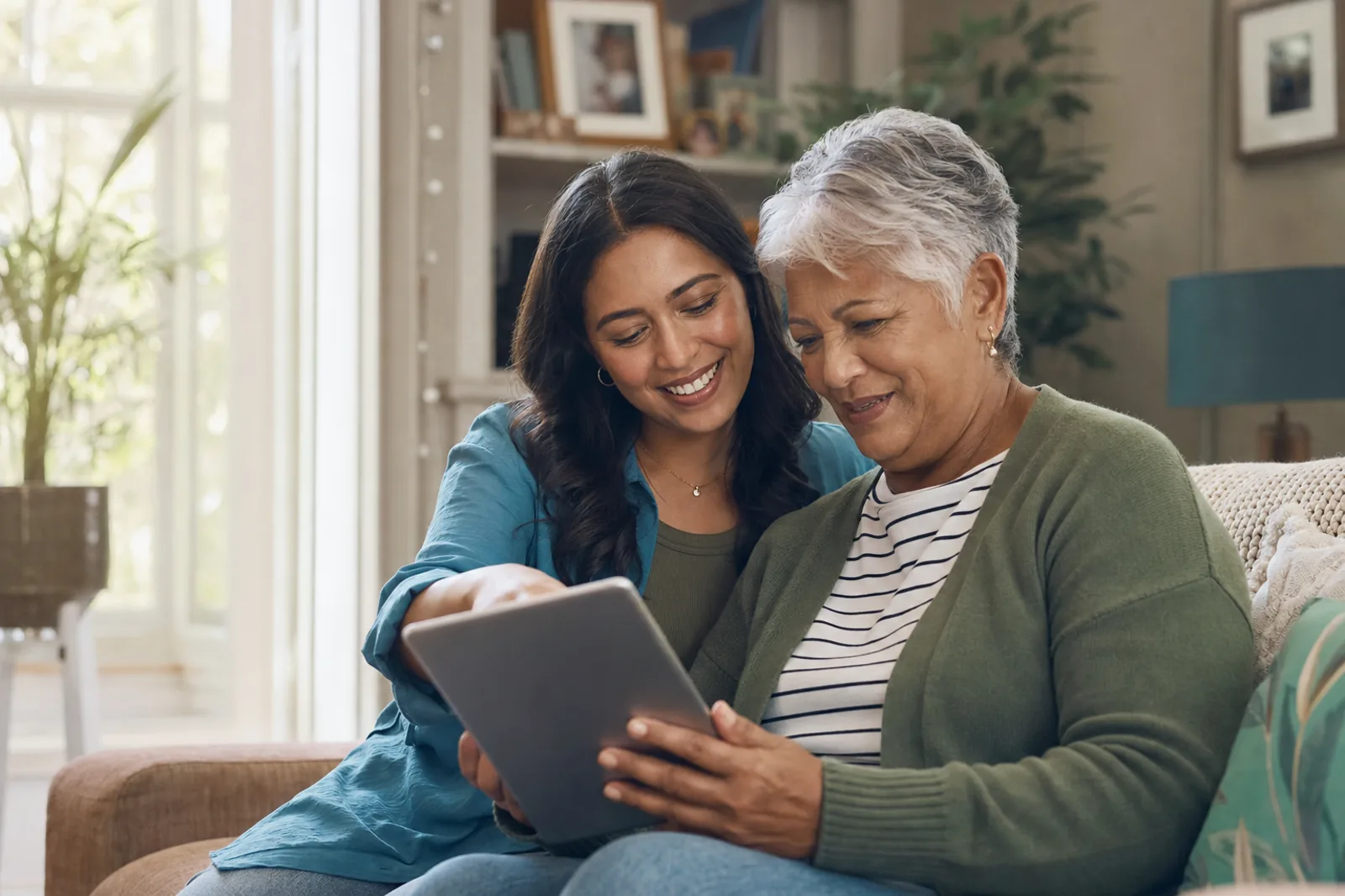 An older adult and a younger family caregiver sitting together at home, looking at a tablet in warm natural light.